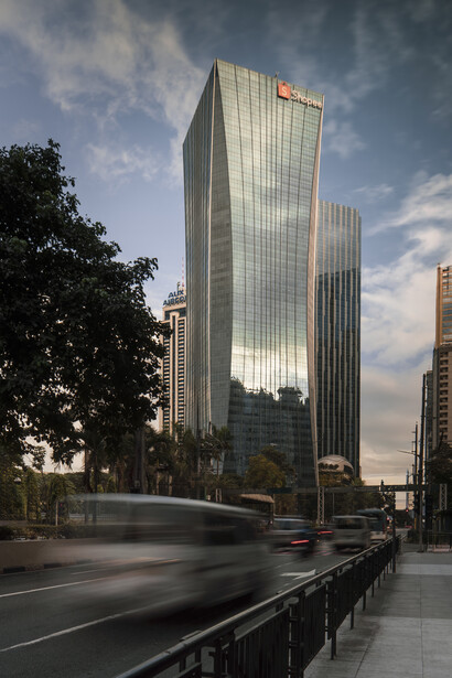 Modern glass skyscraper with reflective facade against cloudy sky, with motion-blurred vehicles and street-level pedestrian barriers.