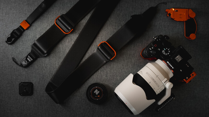 Photography gear flatlay featuring camera body, lens, and black straps with orange accents on gray fabric.