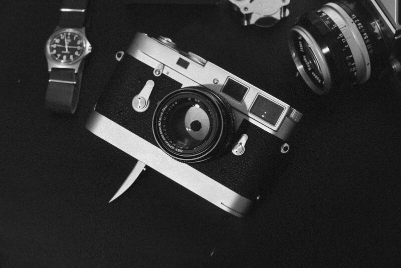 Rangefinder camera with interchangeable lenses, watch, and lens accessories arranged on black background.