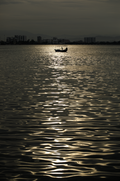Small fishing boat silhouetted against rippling water at dusk or dawn, with distant shoreline and buildings visible.