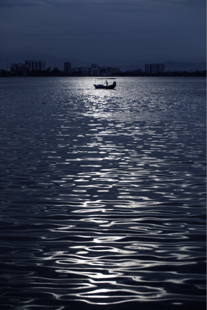 Silhouetted fishing boat on calm water at dusk with city skyline in background.