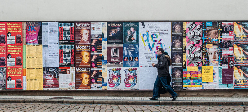 Person in dark clothing walking past a poster-covered wall displaying musical event advertisements and promotional materials.