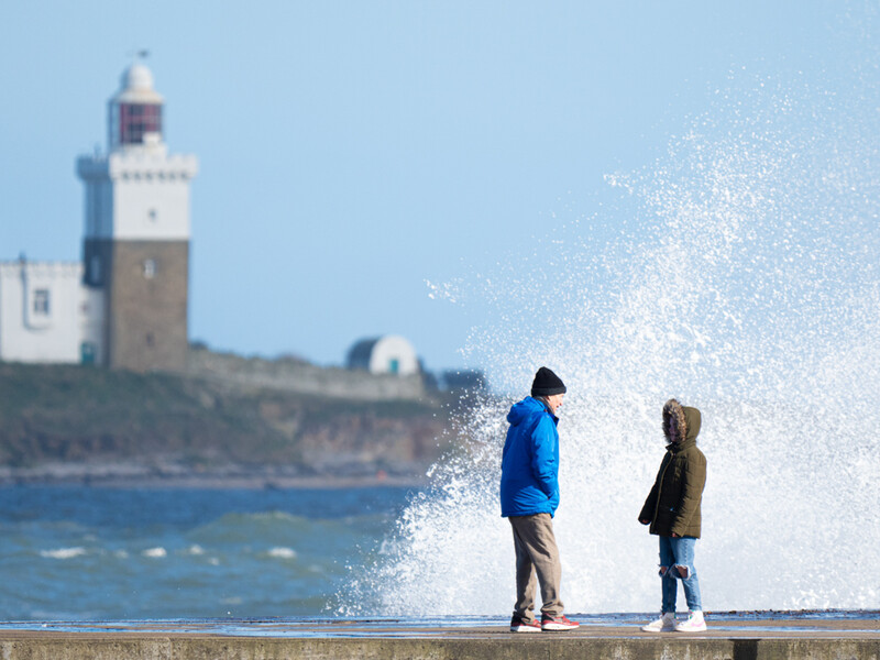 Two people standing on a seawall watch a large wave crash beside them, with a lighthouse in the background.