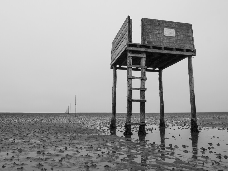 Wooden structure on stilts standing in shallow water and mudflats under overcast sky.