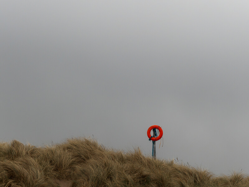 Red life preserver ring mounted on a post above windswept beach grass under overcast sky.