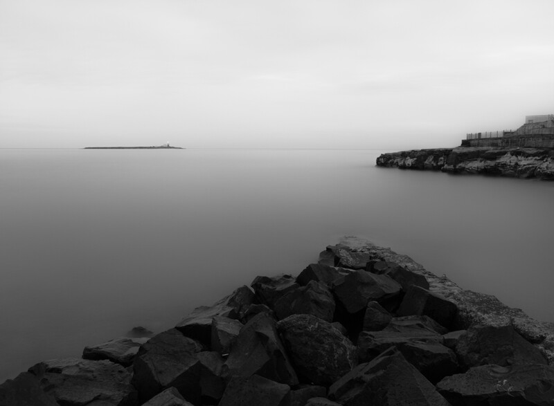 Rocky breakwater extending into still, gray water under overcast sky with distant shoreline.