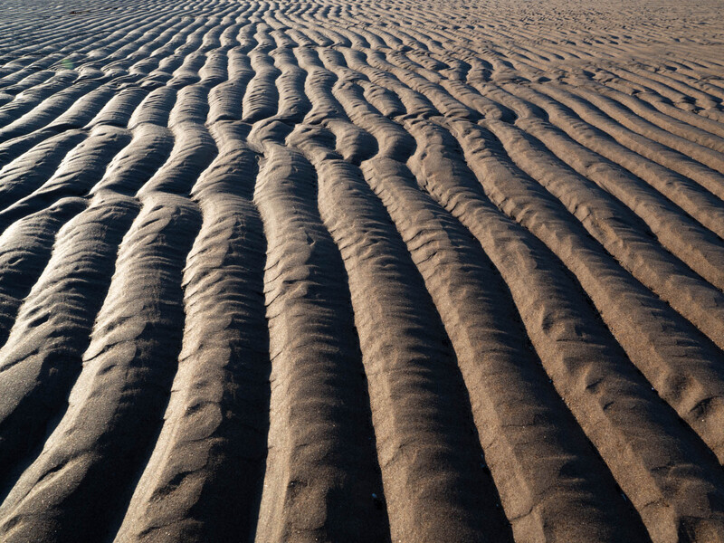 Parallel ripple patterns carved into wet sand, creating deep shadows and rhythmic lines across the beach surface.
