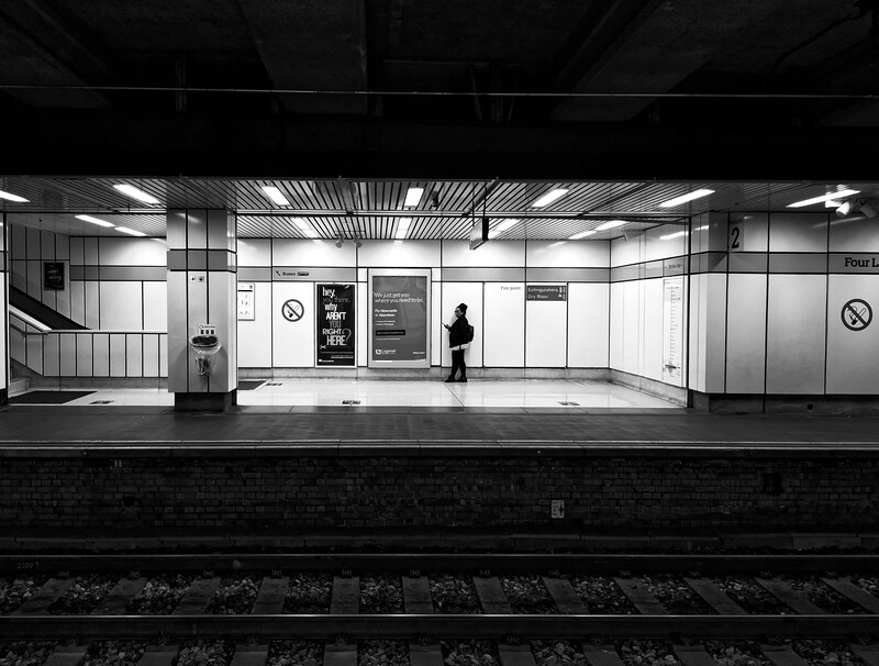 A solitary figure stands on an empty subway platform under fluorescent lighting in black and white.
