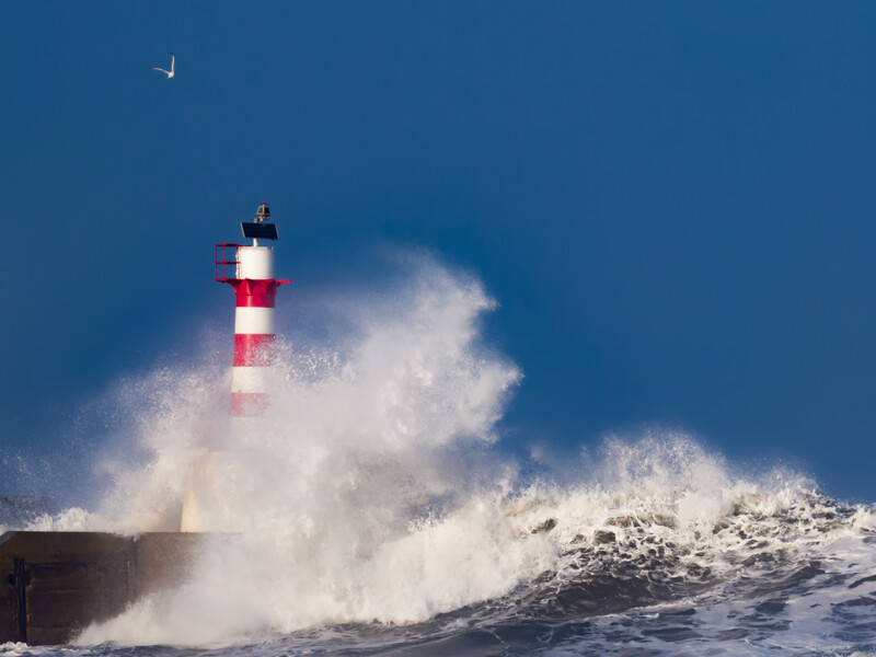Red and white striped lighthouse surrounded by dramatic white ocean waves under clear blue sky.