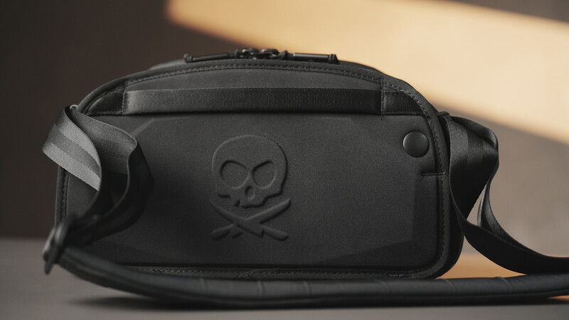 Black camera bag with skull and crossbones emblem and shoulder strap on neutral surface.