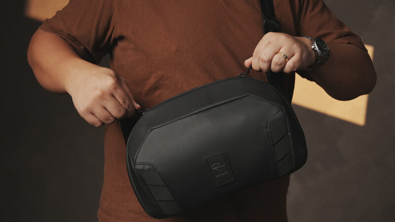 Person in brown shirt holding open a black camera bag with shoulder strap.