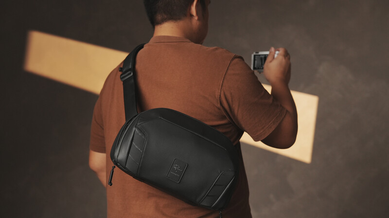 Person wearing brown t-shirt and black shoulder bag holding compact camera against neutral studio background.