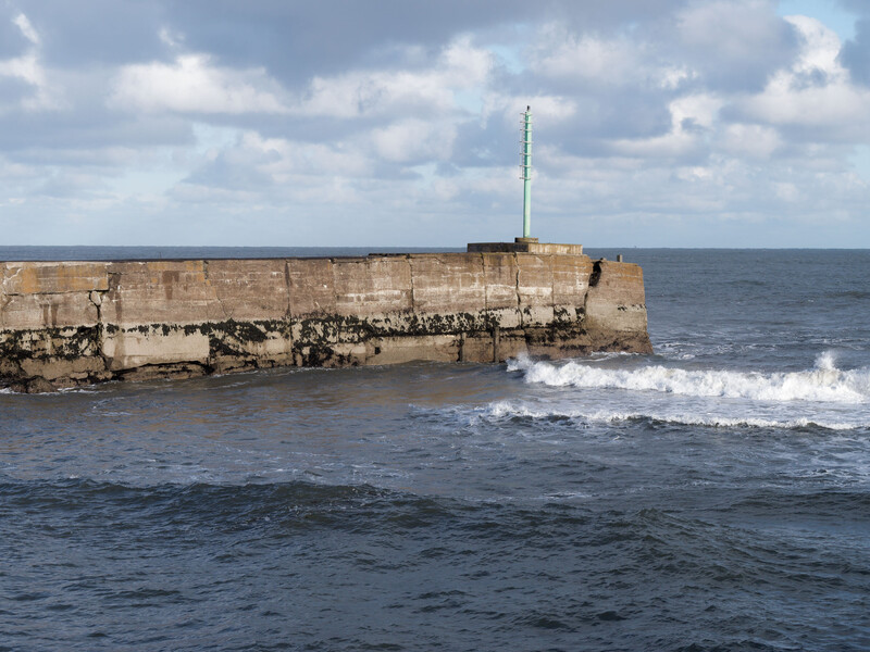 Weathered stone breakwater or pier extending into rough ocean waves under partly cloudy sky.