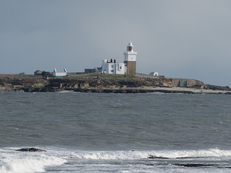 Coastal lighthouse with white keeper's cottage on rocky outcrop surrounded by rough ocean waves.