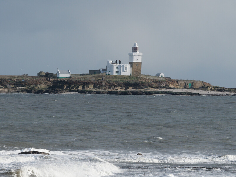 Coastal lighthouse on rocky island surrounded by breaking waves under overcast sky.