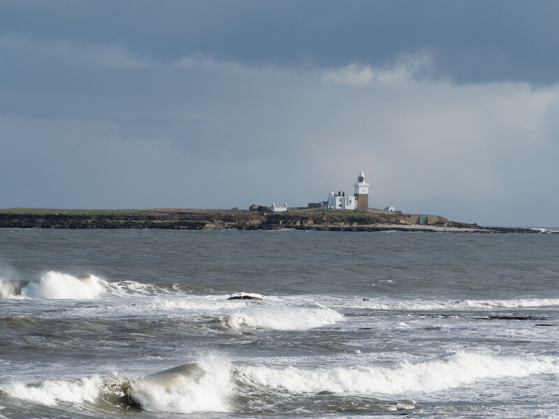 Coastal seascape with breaking waves in the foreground and a lighthouse on a distant rocky peninsula under overcast sky.