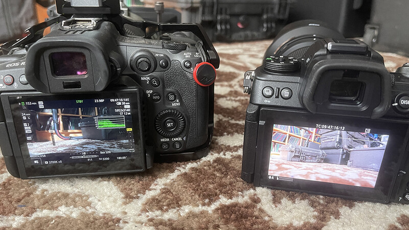 Two professional cinema cameras displayed side-by-side on a patterned carpet, showing their rear LCD screens with video footage.