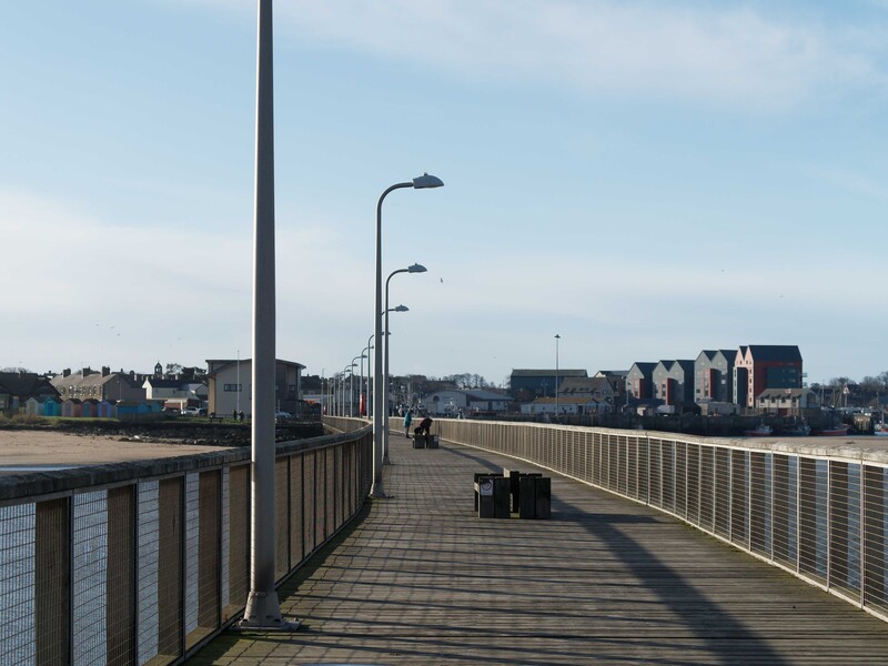 Pedestrian bridge with metal railings extending toward waterfront buildings under overcast sky.