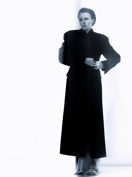 Woman in black coat and dress standing against white background with one hand raised to shoulder.