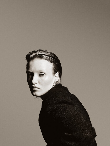 Portrait of a person with styled dark hair, wearing a dark textured sweater, looking over their shoulder against a neutral tan background.