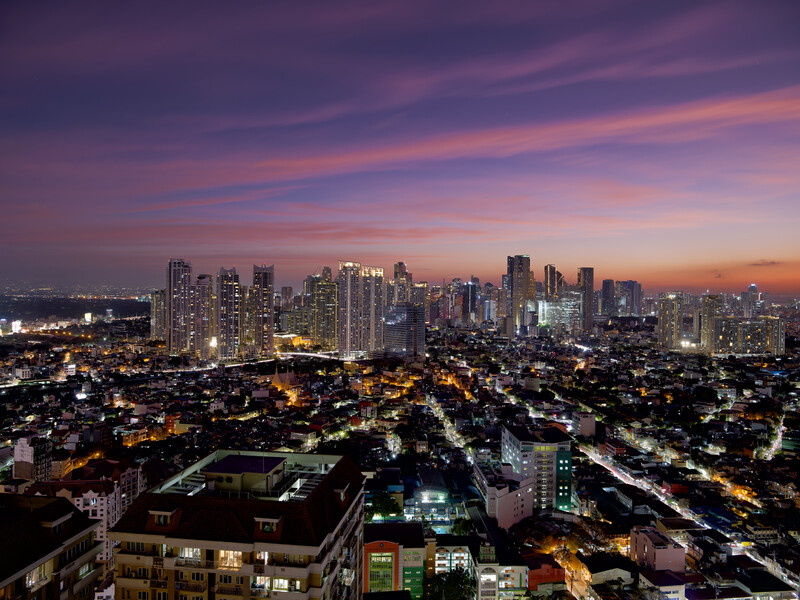 Urban skyline at dusk with illuminated high-rise buildings under a purple and orange twilight sky.