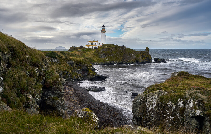 Lighthouse with white buildings perched on rocky headland overlooking turbulent sea.