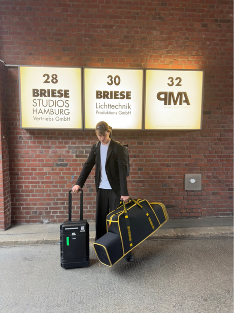 Man in blazer standing with luggage in front of brick wall with illuminated studio signs.