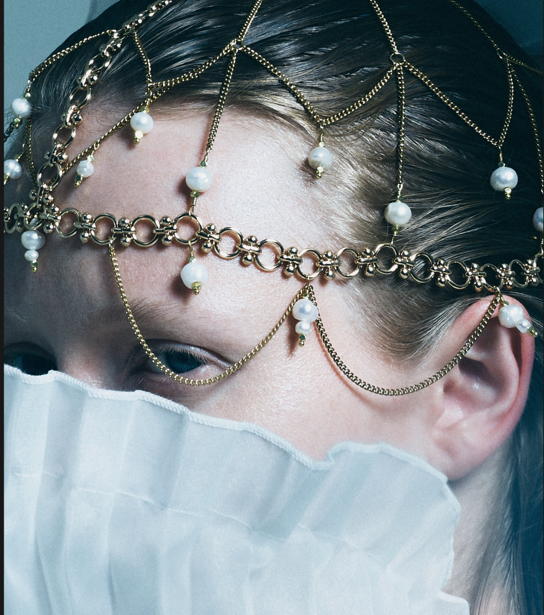 Woman's face framed by layered gold chain jewelry with pearl accents and white tulle fabric.