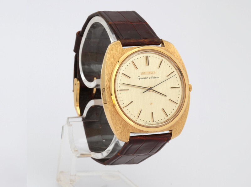 Gold-toned watch with cream dial and dark brown leather strap displayed on a clear stand.