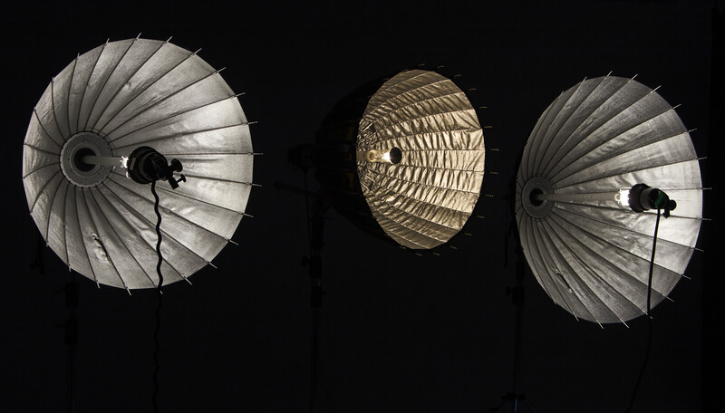 Three studio light modifiers shown against black background: two silver parabolic umbrellas flanking a gold reflective dish.