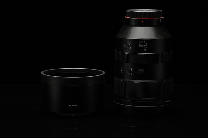A telephoto lens with its lens hood removed, displayed alongside the hood on a dark background.
