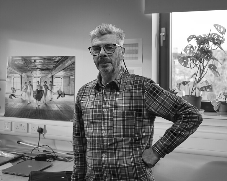 Man wearing glasses and plaid shirt standing in a studio with artwork displayed on walls behind him.