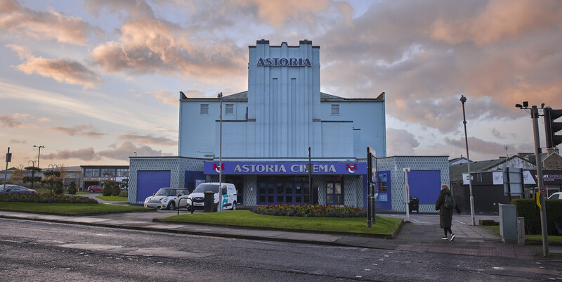 Art deco cinema building with vertical tower element and blue facade under dramatic cloudy sky.