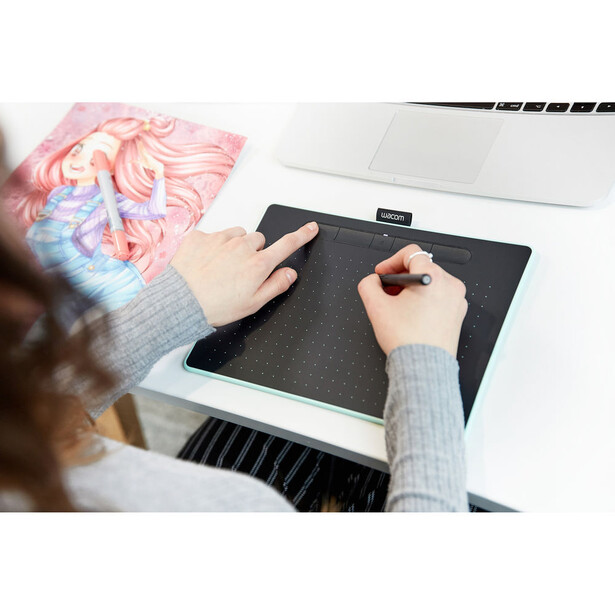 Person's hands using a digital drawing tablet while working at a desk with a laptop and colorful digital art visible.