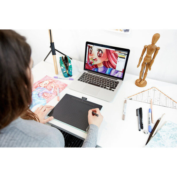 Digital artist working at desk with drawing tablet, laptop displaying illustrated artwork, and creative tools.