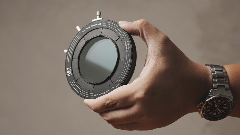 Hand holding a circular lens filter with adjustment knobs against a neutral background.