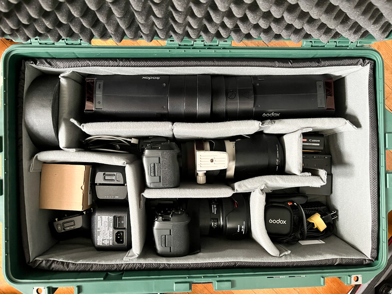 Green hard case with organized photography equipment including lighting gear, flash units, lenses, and accessories neatly arranged with protective foam dividers.