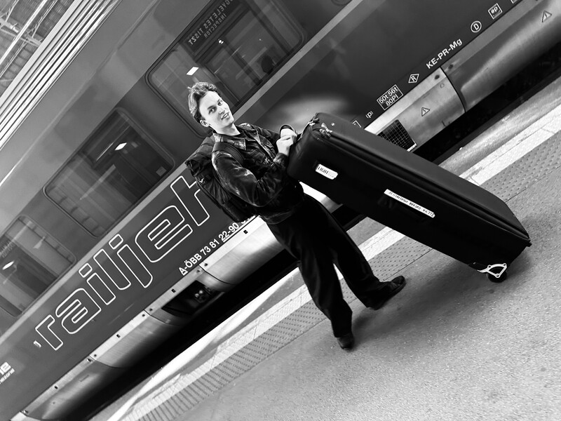 Man in dark suit leaning against a black vehicle on urban street in black and white.