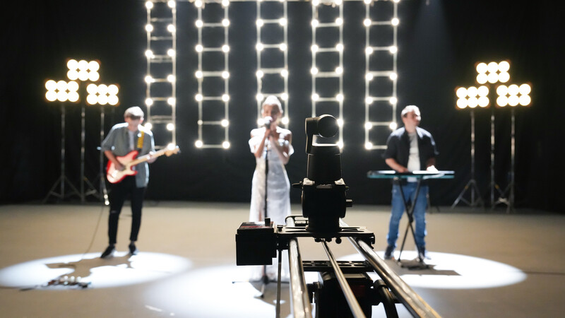 Live band performance with camera on slider rig in foreground, vocalist center stage with guitarist and keyboardist flanking sides under bright LED panel lighting.