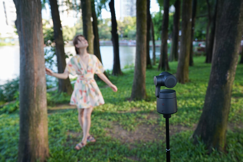 Wireless microphone on stand in foreground with woman in floral dress blurred in wooded park setting.