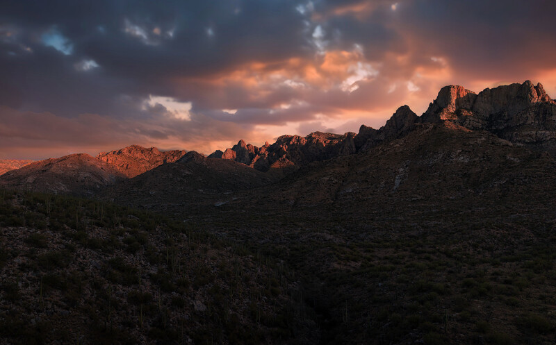 Rocky desert mountains glowing in warm light beneath a dramatic sunset sky with storm clouds.