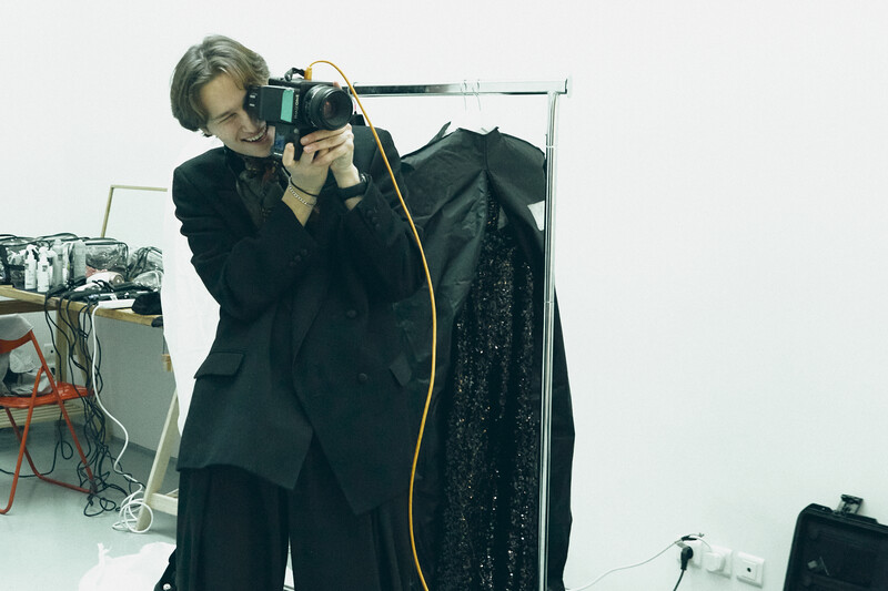 Photographer holding a medium format camera to their face while standing in a studio with black clothing on a rack.
