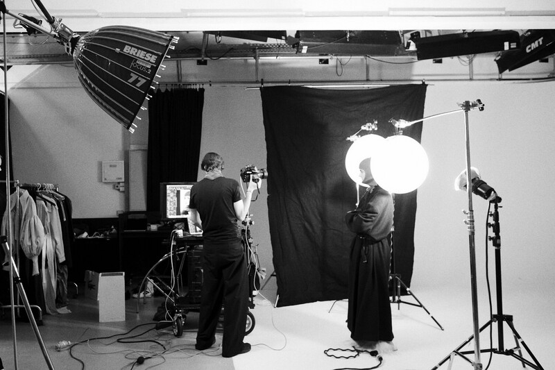 Black and white photograph of a studio portrait setup with two people, professional lighting equipment, and black backdrop.
