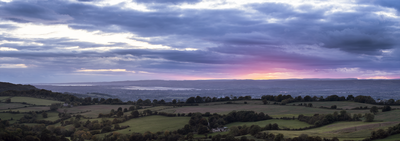 Rolling countryside landscape at dusk with scattered trees, fields, and distant hills under a dramatic cloudy sky with pink and purple tones.