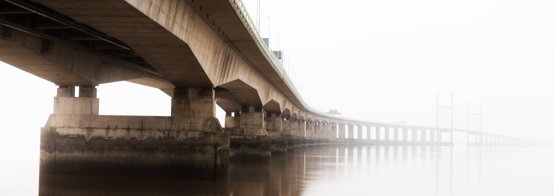 Long concrete bridge with arched supports extending into dense fog over still water.