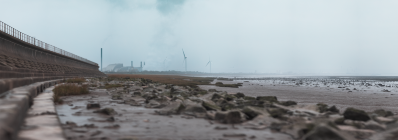 Mudflat landscape with concrete sea wall, industrial structures and wind turbines in distant haze.