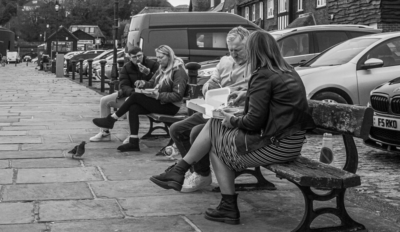 Four people seated on a bench in an urban street, engaged in conversation or looking at documents.