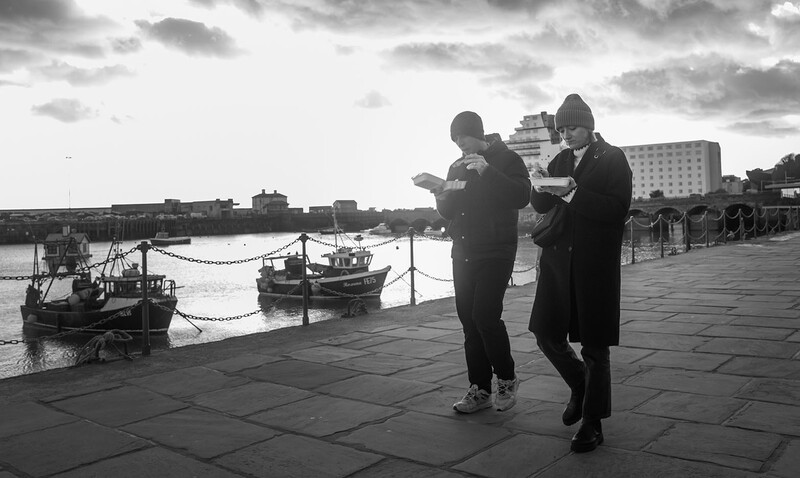 Two people in dark coats check a map while standing on a waterfront promenade beside moored fishing boats.