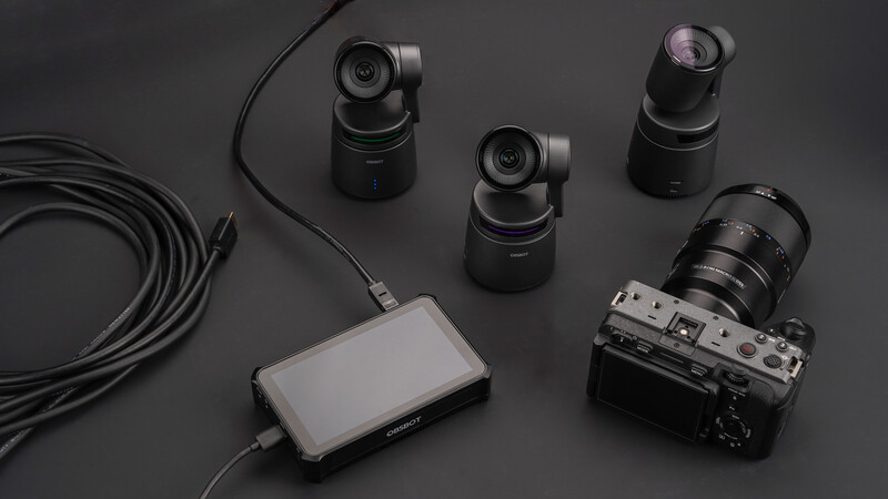 Professional camera equipment laid out on dark gray surface: rangefinder camera with lens, three compact motorized camera heads, external monitor, and coiled cable.