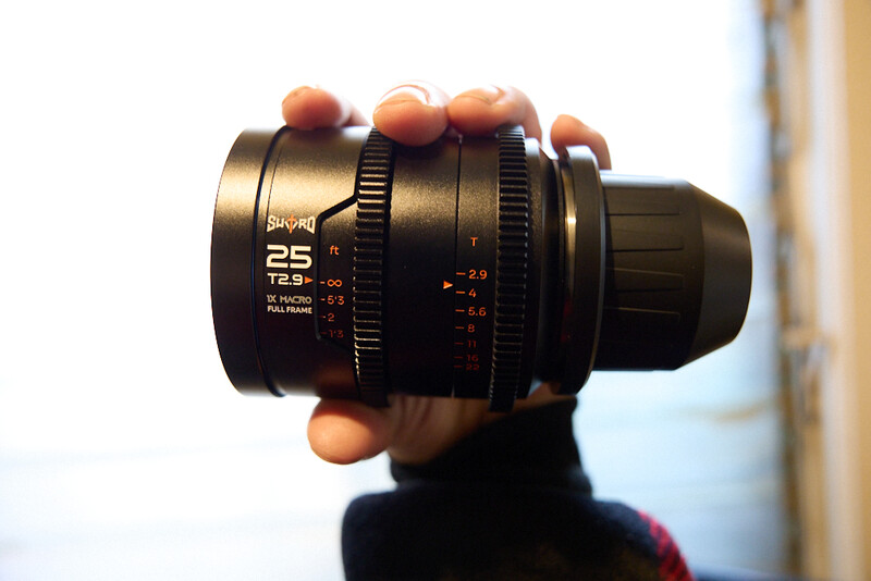A cinema lens with 25mm focal length held up to the camera, showing focus and aperture rings with calibrated markings.